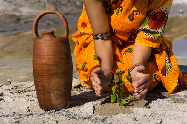 Frau in einem ethnischen Kleid schützt einsamen Sprössling auf trockenem Wüstenboden in der Nähe von Wasserkrug