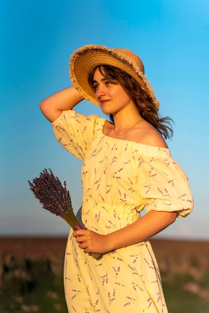 Frau in einem gelben Kleid und einem Hut auf einem Hügel im Sommersonnenuntergang gegen den blauen Himmel