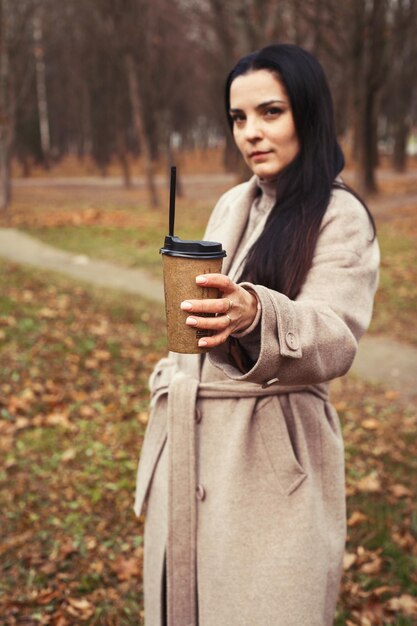Frau in einem grauen Mantel, die mit einer Tasse heißem Getränk im Herbstpark spaziert