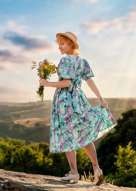 Frau in einem Kleid und einem Hut mit einem Blumenstrauß in ihren Händen auf einem Hügel im Sommersonnenuntergang