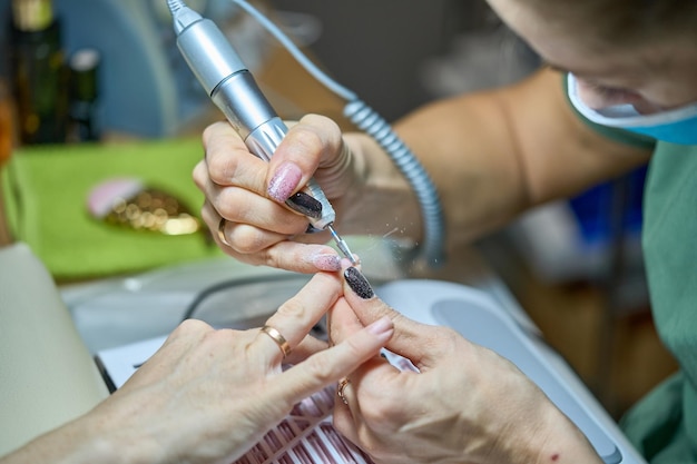 Frau in einem Nagelsalon erhält eine Maniküre in einem Schönheitssalon von einem Kosmetiker mit einer Nagelfile-Bohrmaschine