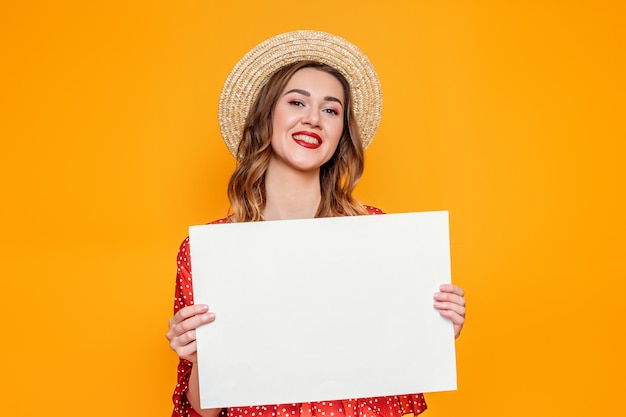 Frau in einem Sommerkleid hält ein A4 leeres Papierplakat und lächelt isoliert über orange Hintergrund