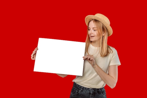 Frau in einem Strohhut und in einem weißen T-Shirt hält weißes leeres Papier, das auf rotem Studiohintergrund mit Beschneidungspfad isoliert ist