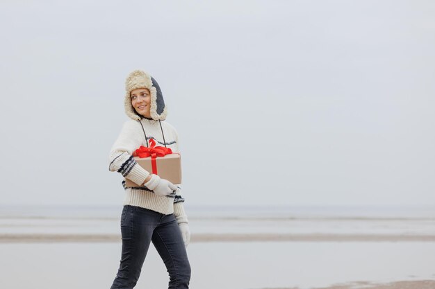 Frau in einem Weihnachtsmann-Hut mit einer Geschenkbox in ihren Händen lächelt eine glückliche Europäerin trägt ein Geschenk für Neujahr oder Weihnachten