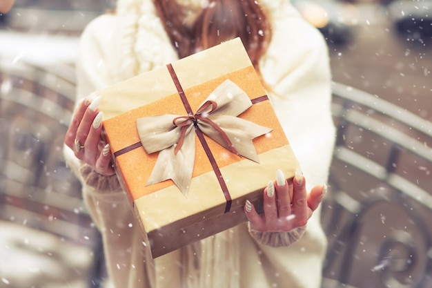 Frau in einem weißen Mantel mit einer großen Geschenkbox mit Seidenschleife auf der Straße bei Schneewetter