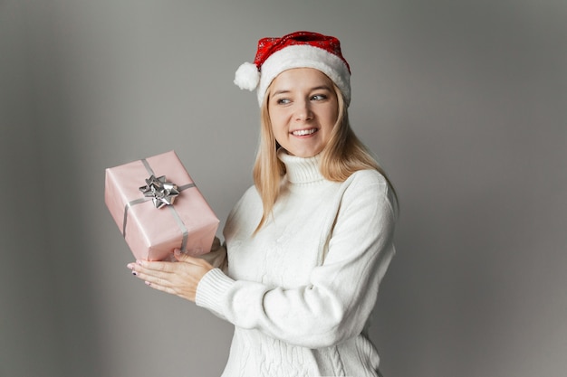 Frau in einem weißen Pullover und einer roten Weihnachtsmütze mit einem rosa Geschenk in den Händen auf grauem Hintergrund