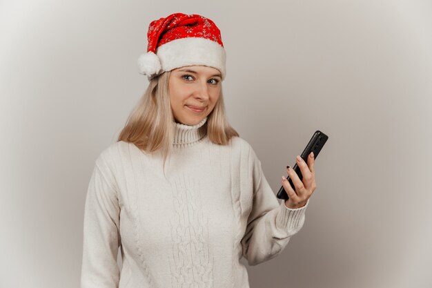 Frau in einem weißen Pullover und einer Weihnachtsmütze mit einem Telefon in den Händen auf einem grauen, isolierten Hintergrund