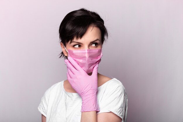 frau in einem weißen t-shirt rosa medizinische maske und handschuhe auf grauem hintergrund