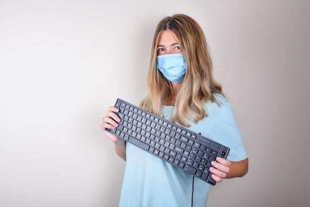 Frau in einer blauen medizinischen Maske und einem blauen T-Shirt hält eine Tastatur in ihren Händen. Zu Hause bleiben. Programmiertag.