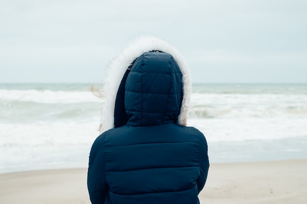 Frau in einer blauen Winterjacke mit einer Haube steht auf dem Winterstrand und betrachtet Meer
