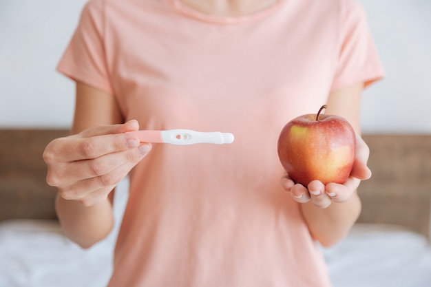 Frau in einer Freizeitkleidung, die einen Schwangerschaftstest und einen Coxs-Apfel als Nährstoffquelle für ihr kleines Baby hält