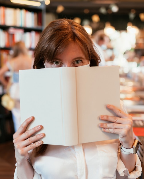 Foto frau in einer medizinischen maske in der bibliothek bedeckt die hälfte ihres gesichts mit einem buch