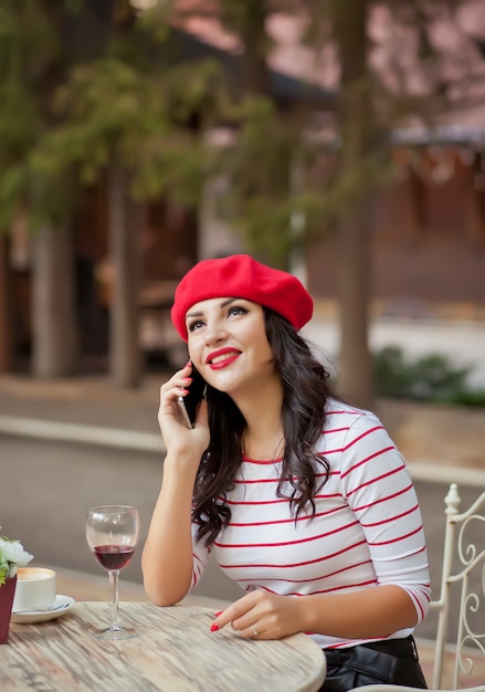 Frau in einer roten Kappe Rotwein im Café im Freien trinkend und am Telefon sprechend