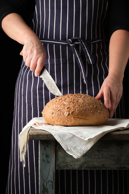 Frau in einer Schürze mit einem Messer in ihrer Hand ungefähr, zum des runden gebackenen Brotes zu schneiden