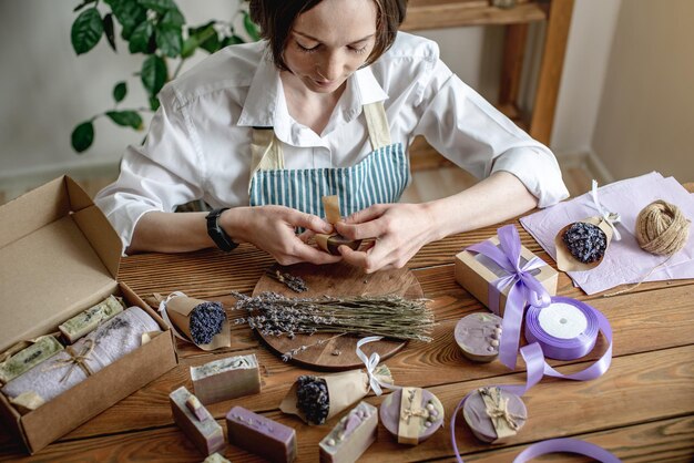 Frau in einer Schürze verpackt natürliche Lavendelseife und dekoriert sie mit Lavendelblüten Konzept von natürlicher Seife und handgefertigten Geschenken