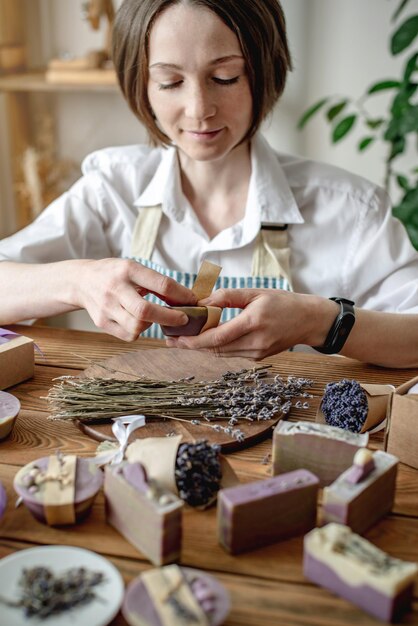 Frau in einer Schürze verpackt natürliche Lavendelseife und dekoriert sie mit Lavendelblüten Konzept von natürlicher Seife und handgefertigten Geschenken