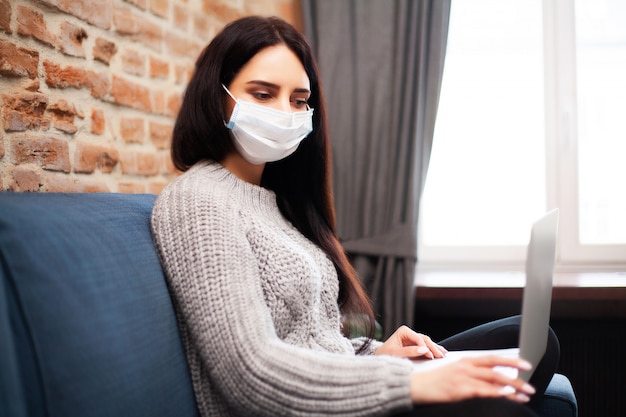 Frau in einer Schutzmaske arbeitet zu Hause auf einem Laptop während einer weltweiten Epidemie eines Virus