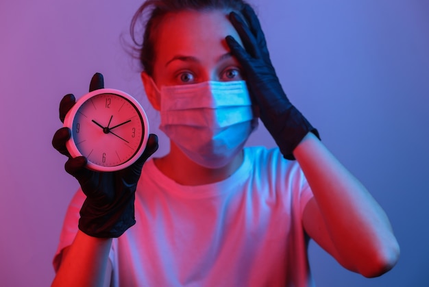 Frau in einer Schutzmaske, Handschuhe hält Uhr. Rot-blaues Neonlicht mit Farbverlauf