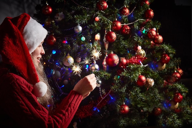 Frau in einer Weihnachtsmütze mit beleuchteten Wunderkerzen auf dem Hintergrund eines Neujahrs- oder Weihnachtsbaums, der mit einer roten Kugel geschmückt ist.