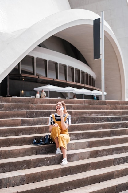 Frau in gelber Latzhose, die auf der Treppe sitzt, lächelnd und im Freien Konzept Lifestyle-Modestil im Freien