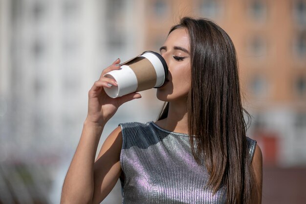 Frau in glänzendem grauem Top mit einer Papptasse Kaffee in der Hand. Brünettes Mädchen mit dunklen Haaren, das Kaffee auf den Straßen der Stadt trinkt. Lässiger Street-Fashion-Look.