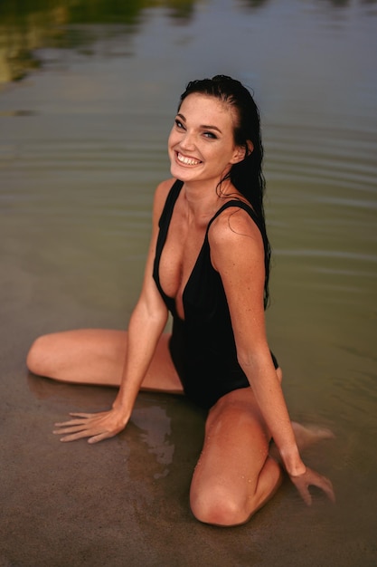 Foto frau in guter laune genießt das leben. fröhliche, glückliche frau im schwarzen akkord-badeanzug sitzt am ufer im wasser.