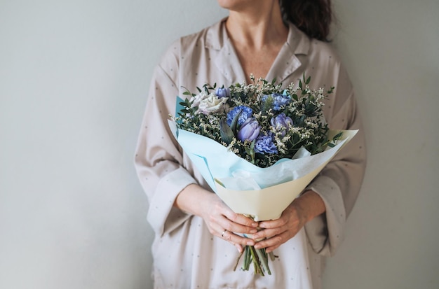 Frau in Hauskleidung mit Blumenstrauß aus blauen und lila Blumen in den Händen auf grauem Hintergrund