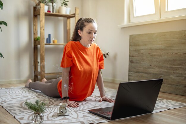 Frau in heller Kleidung auf dem Boden vor einem Laptop macht Yoga mit einer Videostunde