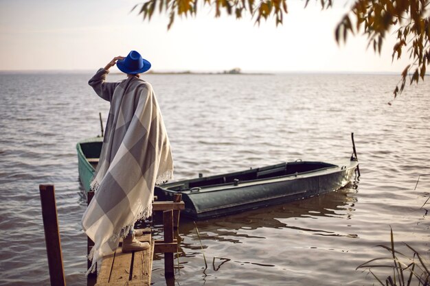 Frau in Jeans und Pullover und Hut steht auf einem Pier mit Booten am See im Herbst