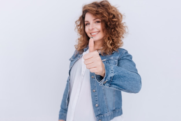 Frau in Jeansjacke, die sich Daumen zeigt. Isolierte graue Wand