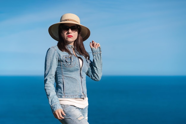 Frau in Jeansjacke, Strohhut und Sonnenbrille, die vor dem Hintergrund des blauen Himmels und des Meeres steht