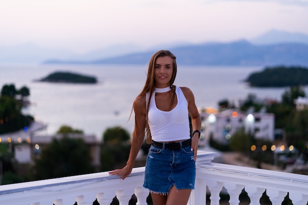 Frau in Jeansshorts, die auf dem Balkon stehen und den Meerblick und den schönen Sonnenuntergang betrachten. Urlaub auf tropischer Insel. Luxus-Lebenskonzept. Entspannen.