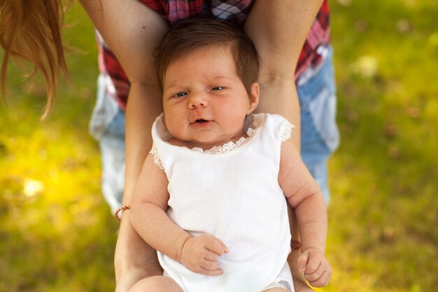 Frau in karierter Bluse, die ein kleines süßes Baby in weißer Kleidung in ihren Händen auf dem grünen Gras hält