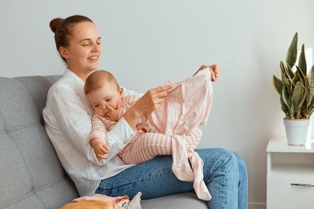 Frau in lässiger Kleidung hat Spaß mit niedlichem Baby, das eine kleine Tochter hält, bewundert neue Kleidung für Kleinkinder und sitzt auf dem Sofa im Wohnzimmer.