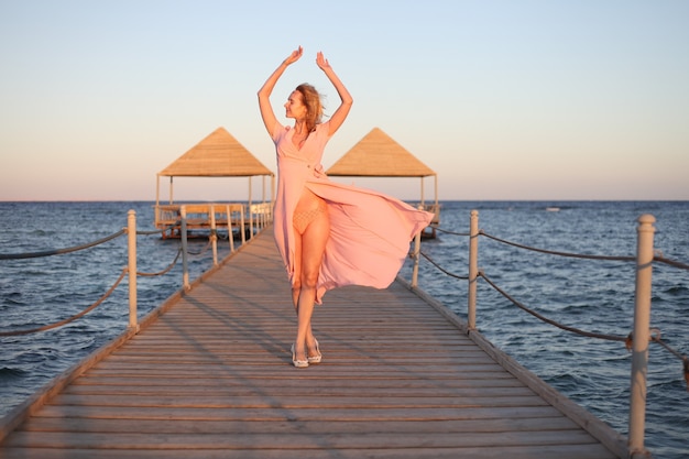 Frau in langem rosa Kleid, die auf Stranddame auf dem hölzernen Hafenferien- und Sommerkonzept geht