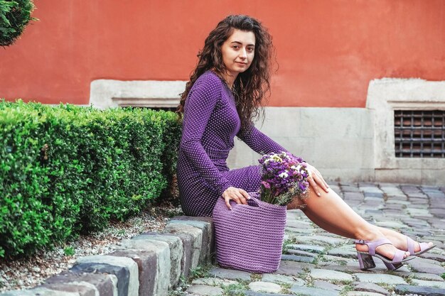 Frau in lila gepunktetem Kleid mit gestrickter Tasche mit Blumen posiert in der Straße der Stadt