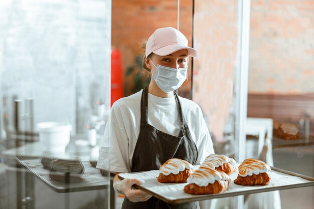 Frau in maske hält tablett mit dekorierten croissants, die in einer handwerksbäckerei stehen