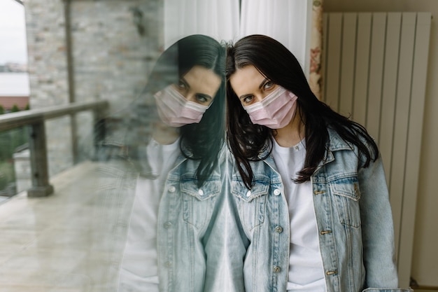 Frau in medizinischer Maske in der Nähe des Fensters Quarantäne zu Hause