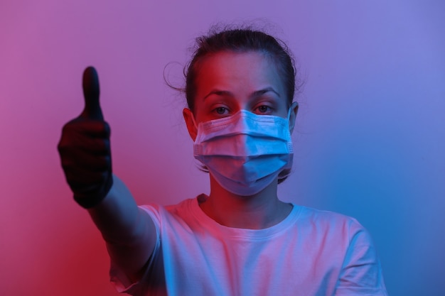 Frau in medizinischer Maske und Handschuhen zeigt Daumen nach oben. Rot-blaues Neonlicht mit Farbverlauf