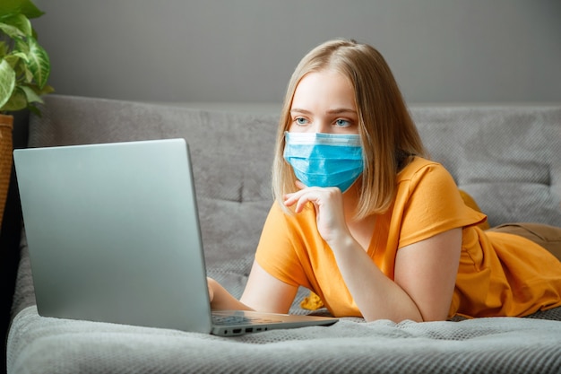 Frau in medizinischer Schutzmaskenarbeit mit Laptop im Home Office liegt auf der Couch. Teenager-Mädchen in Maske Online-Lernausbildung über Laptop-Kovid 19-Sperrzeit. Remote-Arbeit in der Coronavirus-Pandemie.