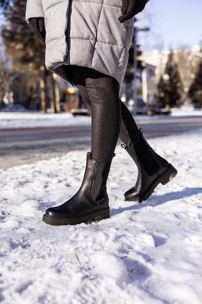 Frau in modischen schwarzen Stiefeln auf weißem Schnee Nahaufnahme Damenbeine in stilvollen Winterlederstiefeln