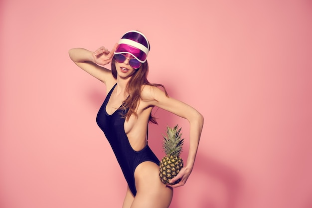 Frau in Plastikmütze mit Visier-Sonnenbrille und Bademode mit Ananas auf Rosa