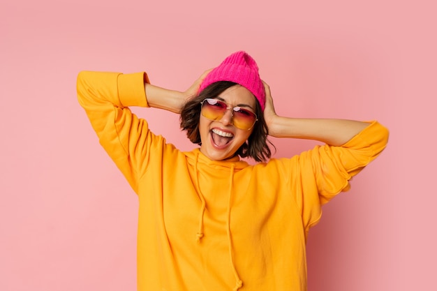 Frau in rosa Hut und orangefarbenem Hoodie mit emotionalem Gesicht, das auf Rosa posiert. Stilvolle Sonnenbrille.