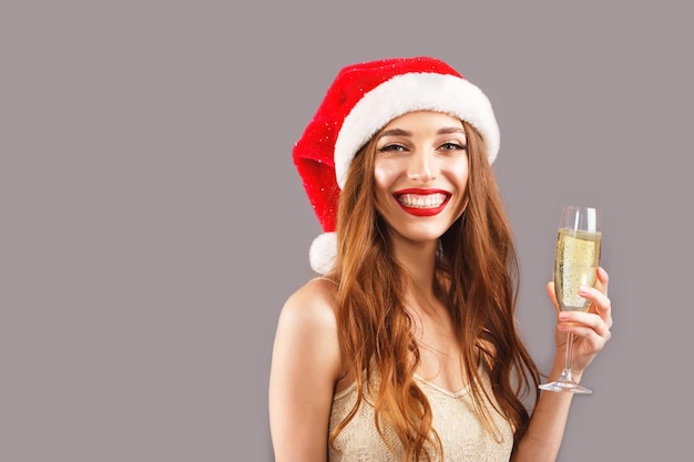 Frau in rotem Weihnachtsmann-Hut mit roten Lippen, die auf dem grauen Hintergrund steht und ein Glas Champagner hält