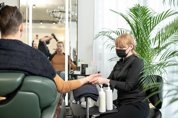 Frau in Schutzmaske massiert dem Kunden in einem Schönheitssalon die Hände