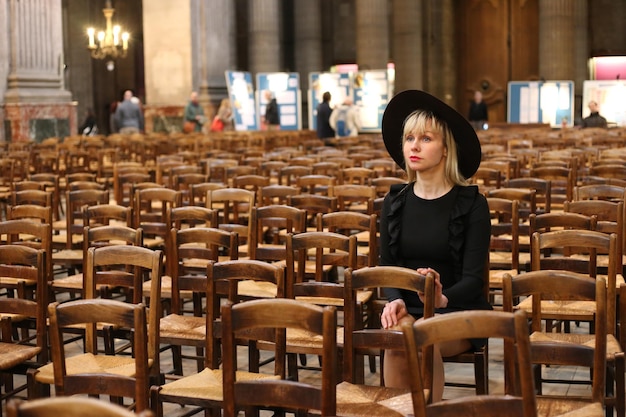 Frau in schwarzem Kleid und Hut sitzt allein in der katholischen Kirche. Gebetszeit