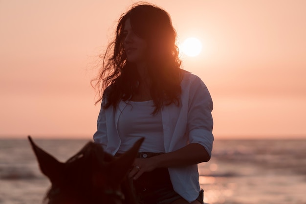 Frau in Sommerkleidung reitet gerne auf einem Pferd an einem wunderschönen Sandstrand bei Sonnenuntergang, selektiver Fokus