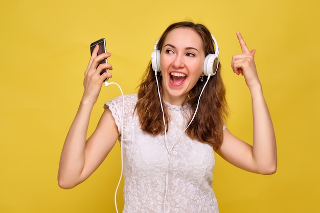 Frau in Sommerkleidung steht auf Gelb, hört Musik in weißen Kopfhörern und schaut in die Kamera des Smartphones, die Selfie macht.
