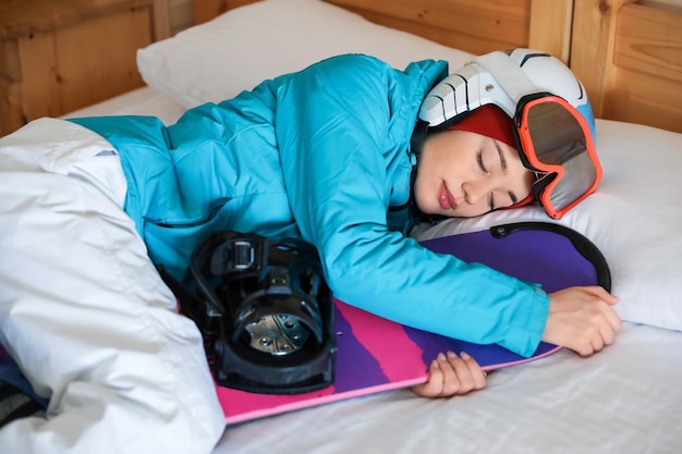 Frau in Sportkleidung mit Snowboard, die auf dem Bett schläft Winterurlaub