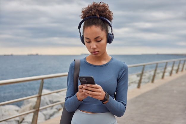 Frau in Spotswear überprüft Musik aus Playlist hält Handy hört Audiospur in Kopfhörern trägt Karemat Spaziergänge am Meer gegen wolkenlosen blauen Himmel